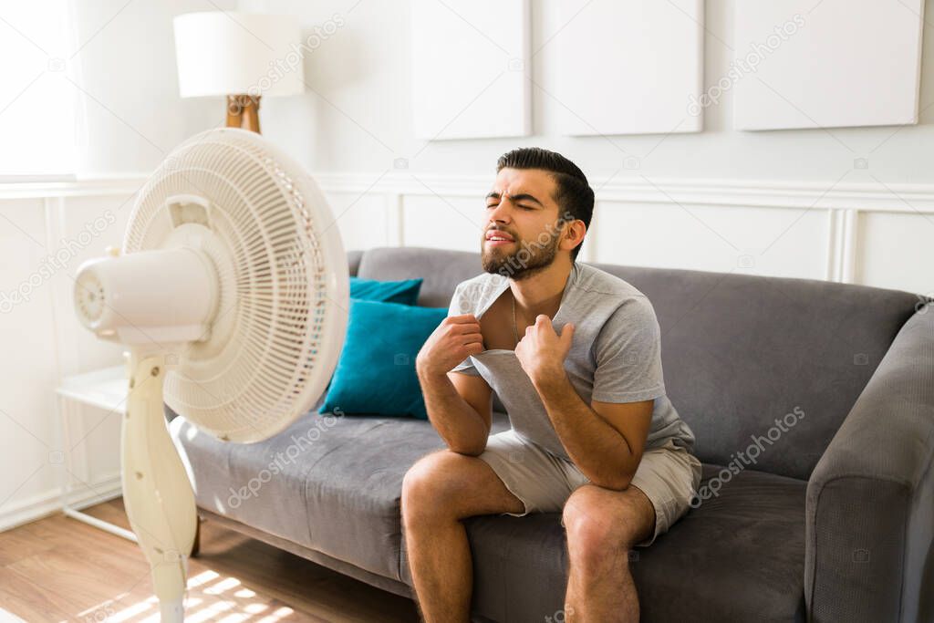 Heat wave. Sweating young man suffering at home because of the hot weather and turning on the fan