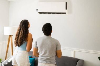 Rear view of a young woman and man enjoying the cold air from the ac unit at home to cool down 