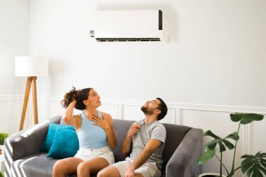 Cheerful young man and woman relaxing on the sofa with the best air conditioner and enjoying the cold air flow 