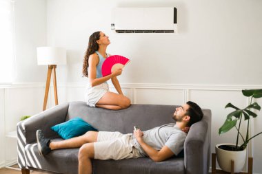 Sad woman feeling hot and trying to cool down sitting below the air in the ac unit with her partner 