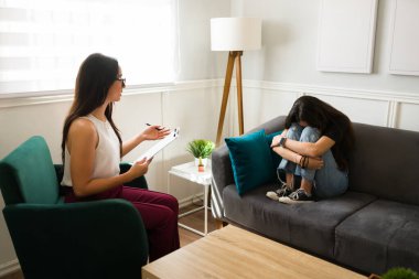 Depresyonla uğraşan üzgün kız seans sırasında kadın terapistini dinlerken dizlerine sarılıp ağlıyor.