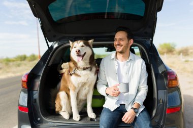 Handsome hispanic man smiling and enjoying the view while sitting in the car trunk with his husky dog