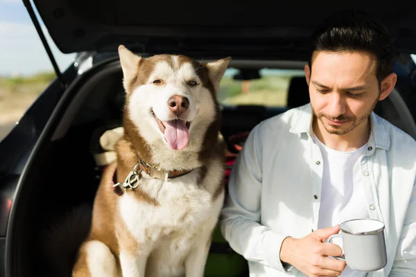 Thoughtful young man drinking coffee and resting while taking a vacation trip by car with his husky dog