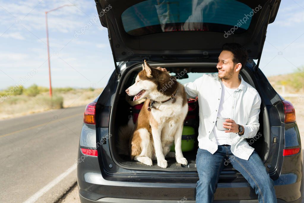 Relaxed young man petting his dog while smiling and resting in his car at the side of the road during a vacation trip