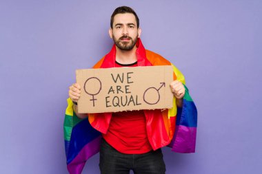Queer young man wrapped in a gay flag and showing a placard with a we are equal message during a march