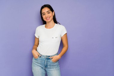 Güzel, kendine güvenen, elleri ceplerinde gülümseyen ve mor bir arka plana karşı poz veren bir kadın.