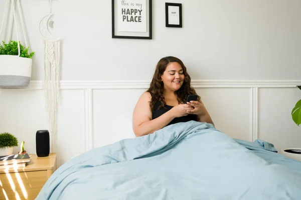 快乐的超重女人躺在被窝里 一边用社交媒体 一边用智能手机发短信 — 图库照片