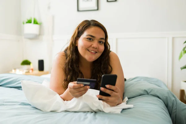 一个微笑的超重女人在床上休息时拿着智能手机和信用卡支付网上购物费用的画像 — 图库照片