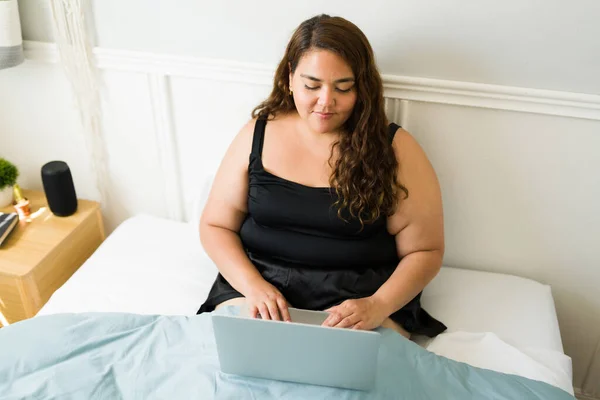 一个穿着睡衣的大块头女人躺在床上一边打字一边在笔记本电脑上工作 — 图库照片