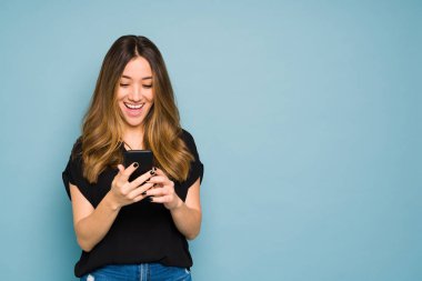 Akıllı telefon tutan genç bir kadın stüdyoda mavi bir arka planda çok heyecanlı görünüyor.