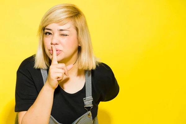Young obese woman with dyed blonde hair winking and putting a finger on her lips to keep quiet in a studio
