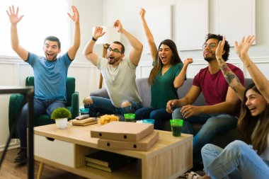 Heyecanlı arkadaşlar televizyon izlerken kollarını kaldırıp bağırıyorlar ve spor zaferini kutluyorlar.