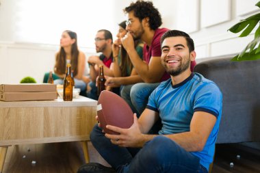 Çekici bir genç adam, gülümsüyor ve arkadaşlarıyla maç izlerken elinde futbol topu tutuyor.