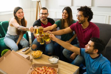 Neşeli çeşitli arkadaşlar birayla kadeh kaldırırken oturma odasında pizzayla toplantının keyfini çıkarıyorlar. 