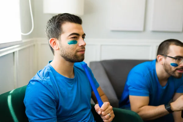 Stressed young man holding a vuvuzela and wearing his soccer team color while watching tv with friends
