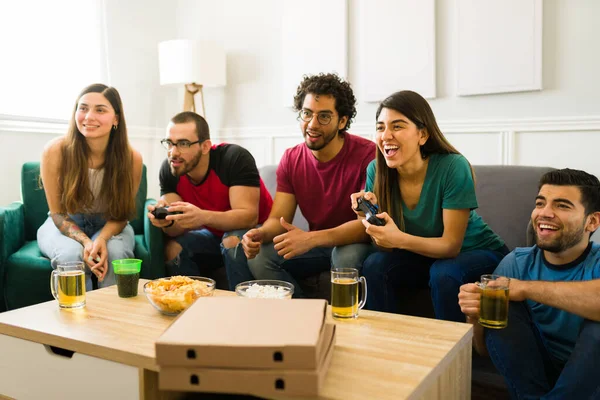 Smiling young women and men laughing while playing video games in the living room while drinking beer