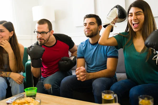 Cheerful sports fans wearing boxing gloves and screaming while watching a box match on tv