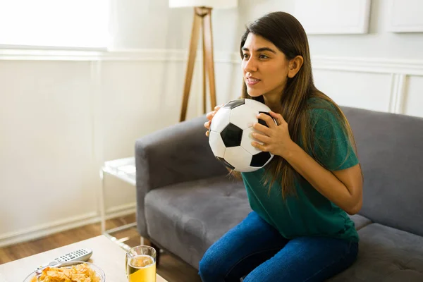 Worried young woman holding a soccer ball and paying a lot of attention to the sports game on tv