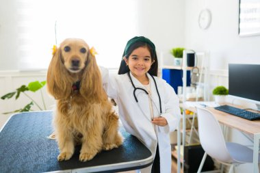 Hayvan hastanesinde cocker spaniel ile veterinercilik oynarken laboratuar önlüklü sevimli genç kız gülümsüyor. 