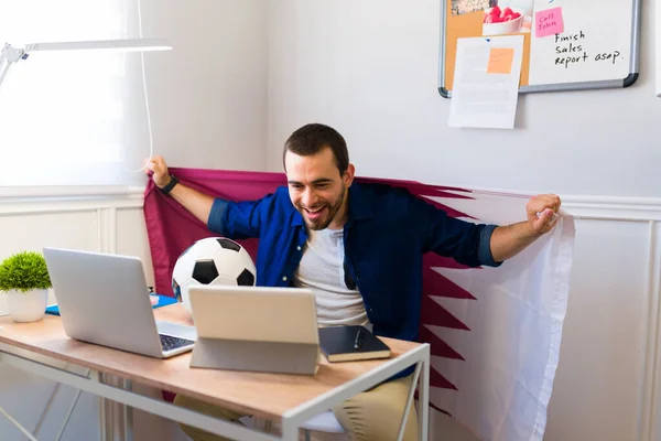 Excited young man using a flag and cheering for his national team while watching the soccer world cup while doing freelance work