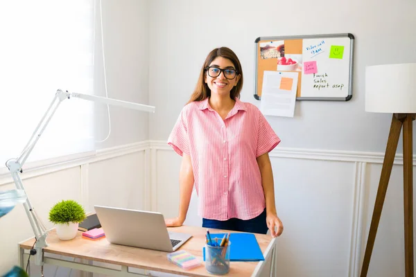 Cheerful hispanic woman wearing glasses and feeling happy while standing behind her work desk for her telecommute job
