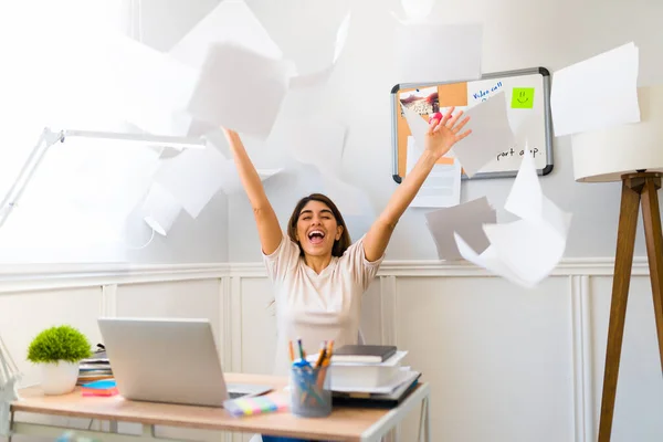 Excited young woman throwing papers and celebrating finishing her work while sitting at her desk at home