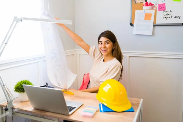 Cheerful professional woman smiling while holding the construction plans and working as an architect in the office