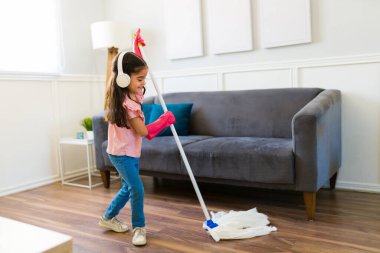 Cheerful young girl with headphones using a broom and cleaning the floor while doing housework