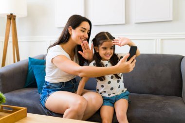 Neşeli anne ve küçük kızı akıllı telefon kullanırken selam veriyorlar ve evde aileleriyle görüntülü görüşme yapıyorlar.