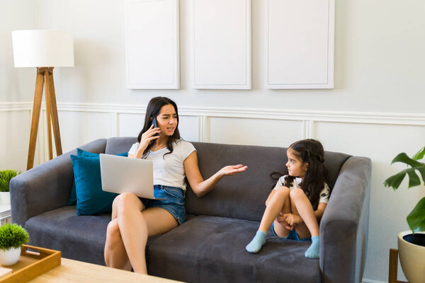 Angry latin mom working and talking on the phone while scolding her young daughter for bad behavior 