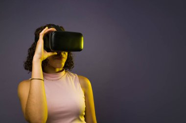 Attractive woman putting on virtual reality glasses and using immersive technology standing in front of a dark background with copy space