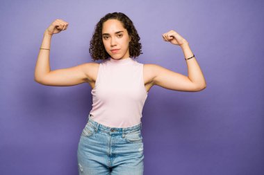 Güçlü, çekici bir kadın kendini çok güçlü hissediyor ve mor bir arka plan önünde kadınları güçlendirmek için kollarını gösteriyor. 