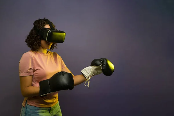 Young brunette woman using boxing gloves and watching a box match simulation or video game with virtual reality glasses