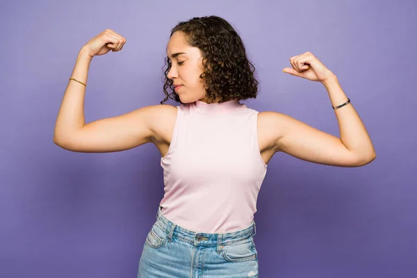 Brunette empowered woman with curly hair looking at her strong arm biceps and showing her strength on a studio background