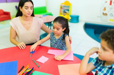 Anaokulu öğretmeniyle anaokulunda kesim yaparken makas kullanan küçük Latin bir kız.