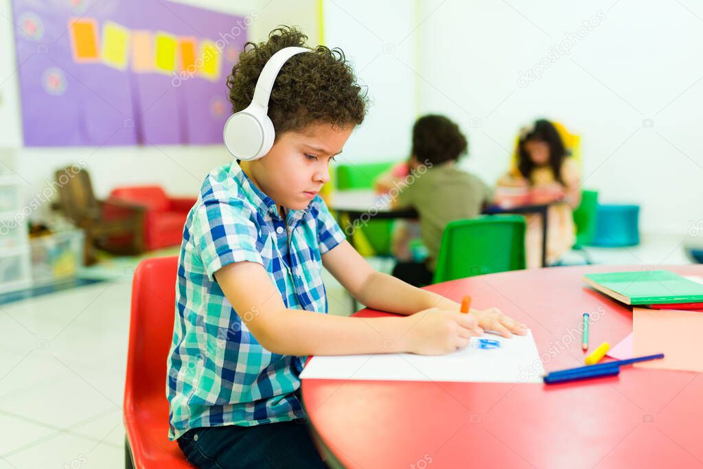 Sad preschool student boy with autism wearing headphones and feeling isolated from his classmates in kindergarten 