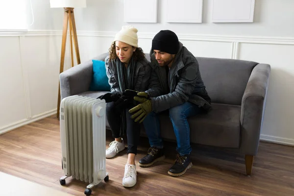 Sad couple wearing winter clothes and sitting in front of the heater in the living room because of the cold weather