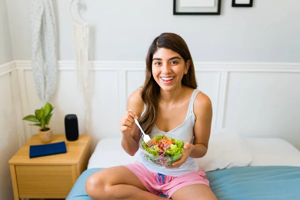 Excited attractive woman looking at the camera while eating a delicious green salad while sitting in bed