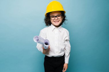 Happy little boy with glasses and a yellow helmet using construction plans while playing architect against a blue background