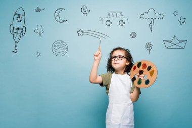 Smart artistic boy using his imagination while painting and using a paintbrush against a blue background with copy space