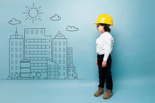 Adorable kid with a engineer aspiring career playing to work in construction next to buildings graphics