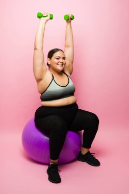Excited obese woman smiling and making eye contact while sitting on a fitness ball lifting dumbbell weights during her exercises