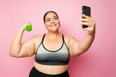 Beautiful fat young woman taking a selfie lifting dumbbell weights with her smartphone while doing her fitness workout