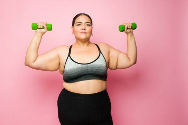 Obese hispanic woman looking at the camera while lifting dumbbell weights and exercising to lose some weight