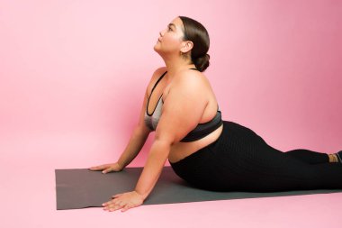 Side view of an active fat sporty woman doing an upward dog pose while exercising with yoga against a pink background
