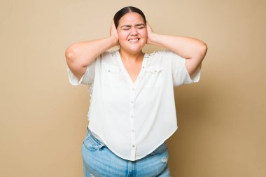 Overwhelmed fat woman feeling anxious because of the loud noise while covering her ears against a yellow background