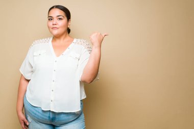 Confident determined plus size woman in casual clothes looking serious pointing to copy space on a yellow background
