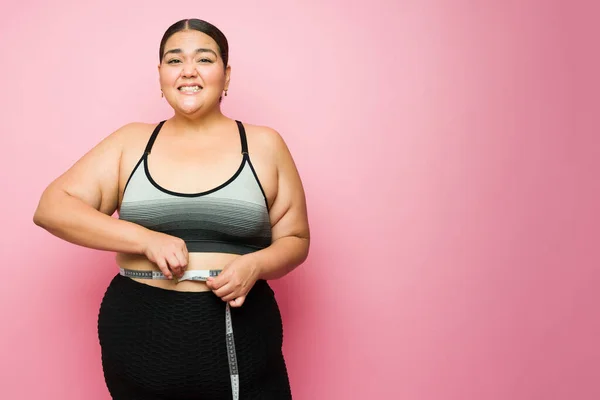Upset obese woman feeling worried and stressed about her body weight while using a tape measure in front of a pink background with copy space