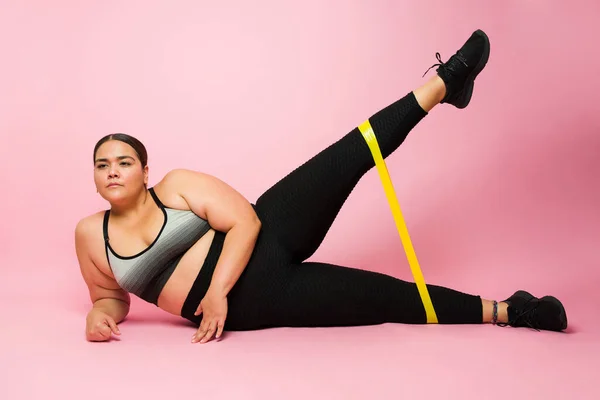 Determined obese woman working out and doing side planks using a stretching fitness band in front of a pink background