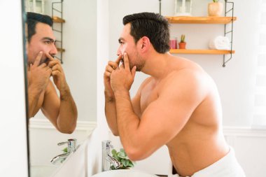 Profile of a strong latin shirtless man with acne looking in the bathroom mirror squeezing a pimple while grooming